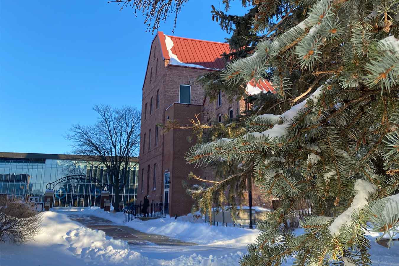 Campus photo in the snow featuring the campus archway, East Hall, and an evergreen tree